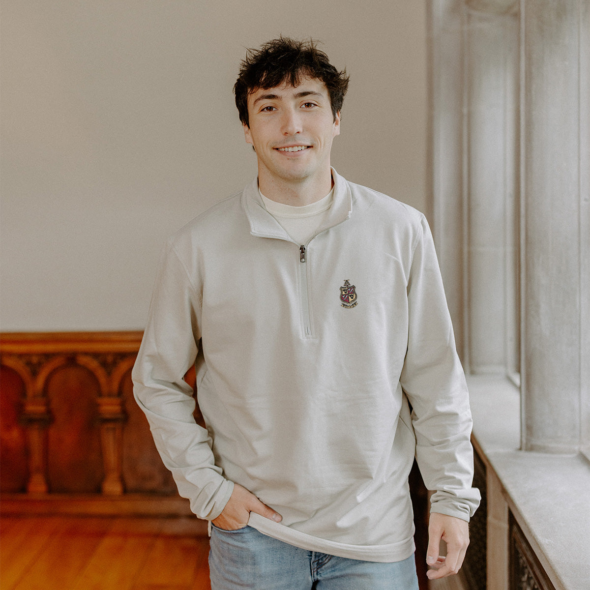 Male model wearing the smoke-colored Delta Chi Embroidered Luxe Quarter Zip with crest embroidery, standing by a window in a formal setting.