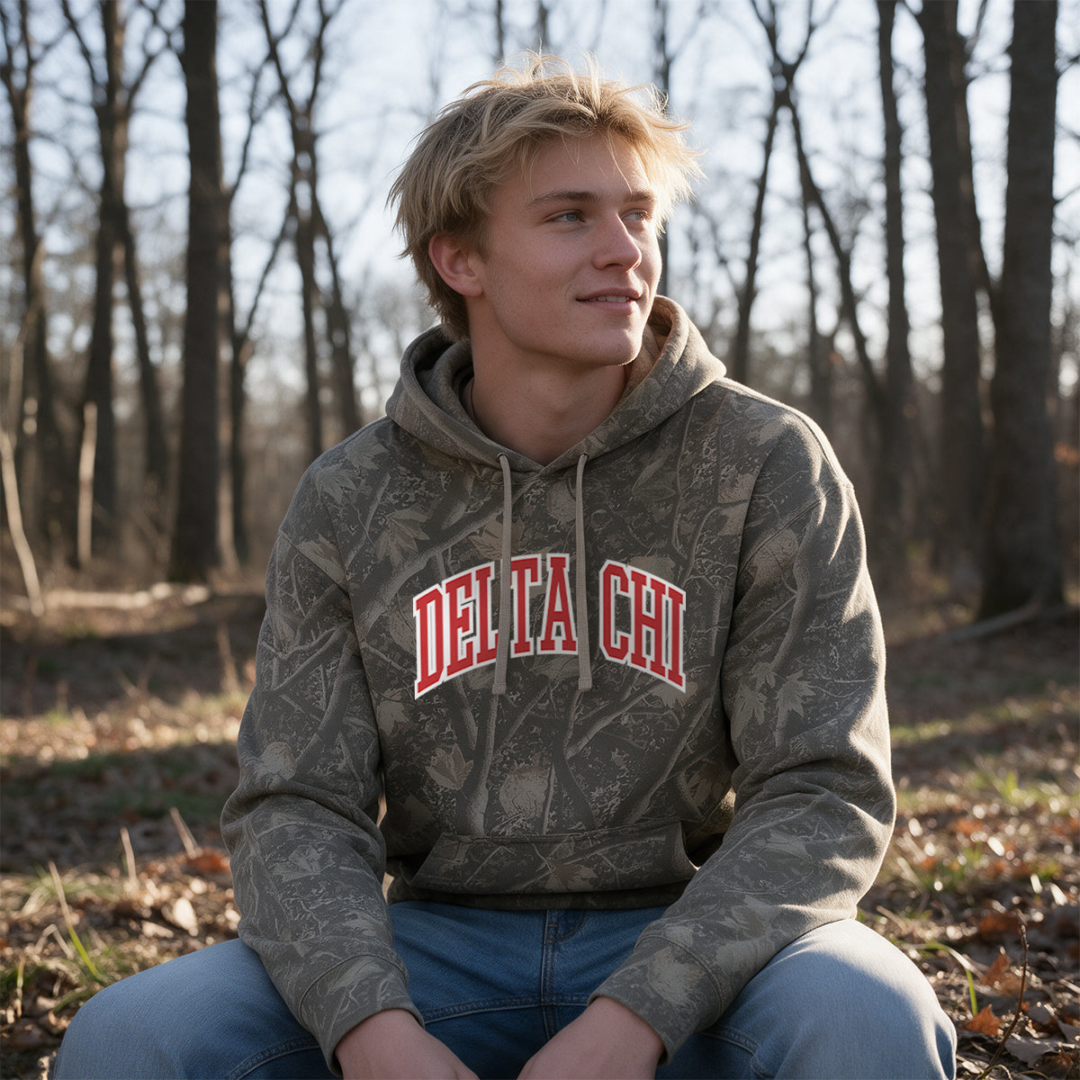 College-aged male wearing the fraternity Deadwood Camo Hoodie with red and white Delta Chi lettering, seated outdoors in a wooded setting.