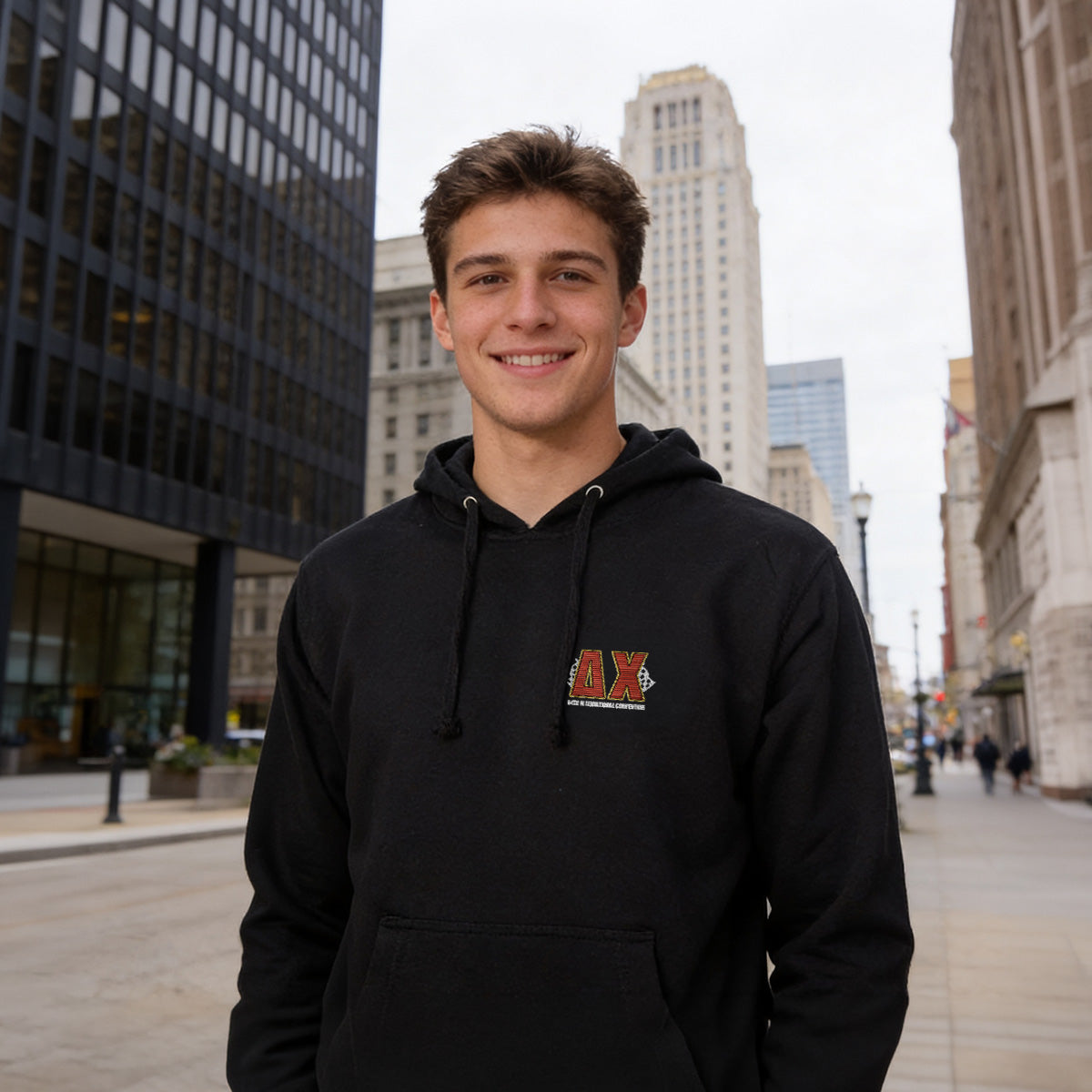Young man wearing a black Delta Chi hoodie with a small embroidered chest graphic, standing in a city street setting.